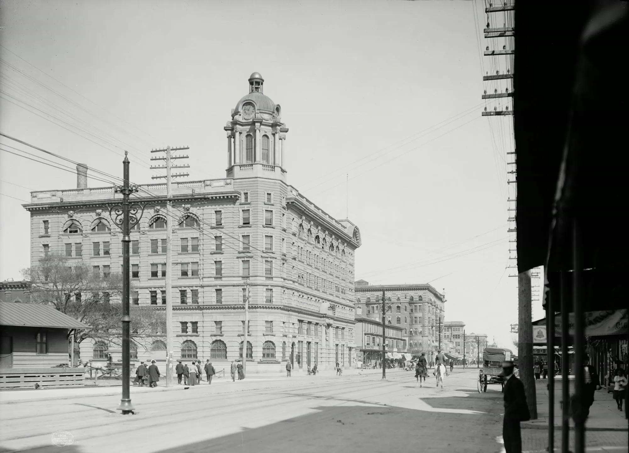 A black-and-white photograph of Atlantic Avenue: a tall stone building with a domed clock tower on the corner, people on the sidewalk, and riders on horseback in the street.