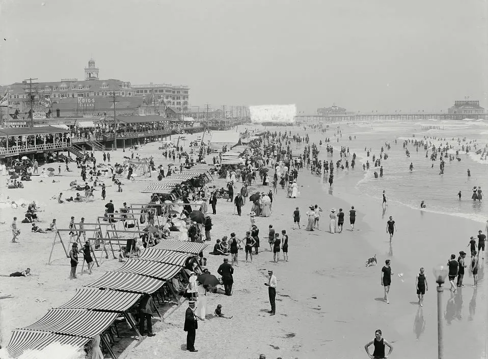 A black-and-white photograph from high on the Boardwalk: a crowded beach with striped awnings, bathers in the surf, hotels along the shore, and piers in the distance.