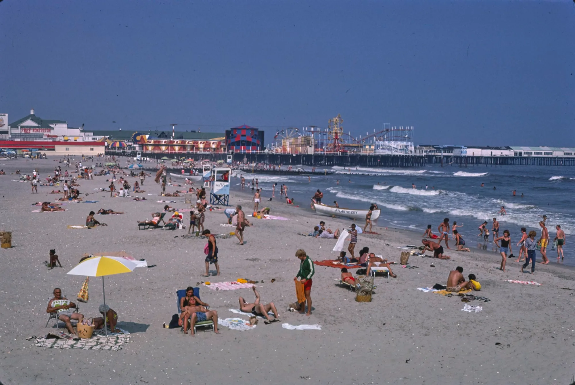 A crowded summer beach in colour: umbrellas, towels, and a lifeguard stand, with the amusement piers and their rides along the water behind.