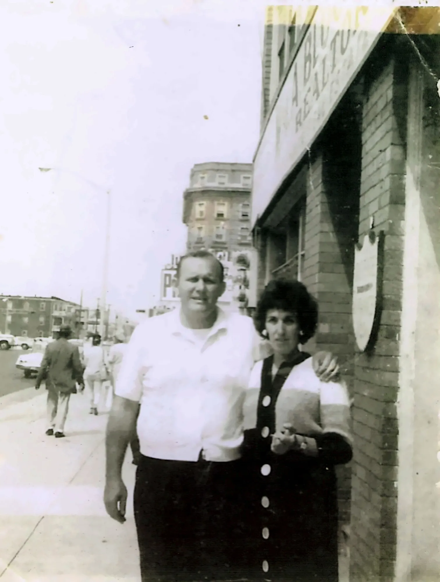An old black-and-white photograph, warm with age: a man in a white short-sleeved shirt with his arm around a woman in a striped cardigan, on the sidewalk outside a brick office with a Bloom Realtors sign above.