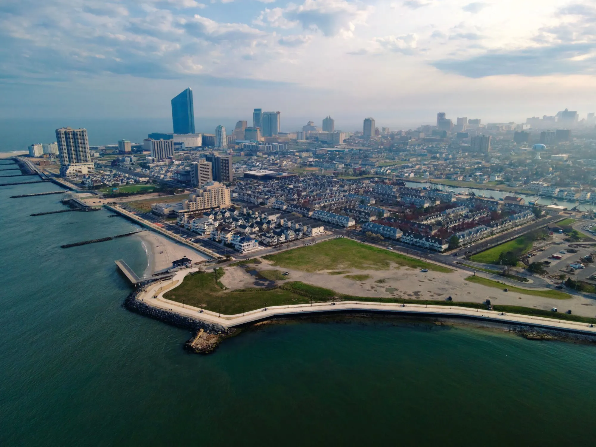 Atlantic City from the air over the inlet: a seawall promenade and park in the foreground, rows of houses and the marina behind, and the casino towers along the shore under light clouds.